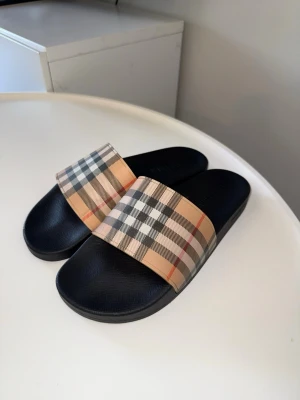 Burberry slides - Säljer ett par Burberry slides i storlek 46. De är i bra skick och har endast mindre tecken på användning. Väldigt bekväma och perfekta för sommaren, semestern eller till vardags.  Klassisk Burberry-design med den ikoniska loggan. Skorna är väl omhändertagna och fortfarande i fint skick. Se bilderna för exakt skick.  Storlek: 46   Märke: Burberry   Skick: Bra  Hör gärna av dig om du vill ha fler bilder eller mer information.