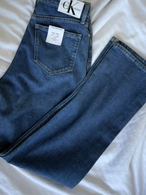 Blå high waist jeans Calvin Klein -  Helt nya! Snygga blå high waist jeans från Calvin Klein Jeans. Klassisk femficksmodell med raka ben och tydliga sömmar. Tillverkade i mjukt denimtyg som ger en avslappnad men ändå stilren look. Perfekta för dig(W26/L30) som gillar tidlös och enkel stil. Storlek S