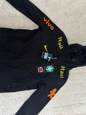 Svart Nike Brasil jacka med huva - Snygg svart träningsjacka från Nike med Brasilien CBF-logga, gula och orangea tryck samt sponsorer. Jackan har huva, dragkedja och är tillverkad i lätt, ventilerande material med Dri-FIT teknologi. Perfekt för fotbollsträning eller sportig stil. Aldrig använd 