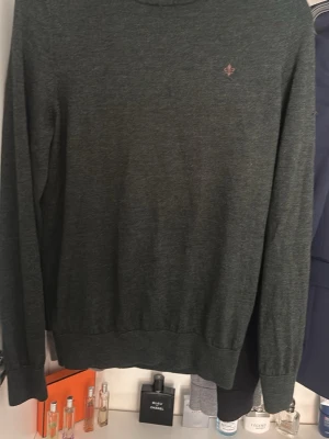 Mörkgrön stickad tröja - Mörkgrön stickad tröja med rund halsringning och diskret broderad logga på bröstet. Tröjan har långa ärmar och ribbade muddar vid ärmslut och nederkant. Perfekt för lager på lager och enkel att matcha.