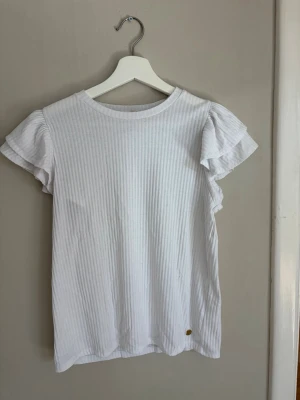Vit ribbad topp med volangärm - Säljer en söt vit ribbad topp med volangärmar och rund hals. Toppen har en liten dekorativ knapp med ananasdetalj längst ner. Perfekt till jeans eller kjol för en fräsch look.