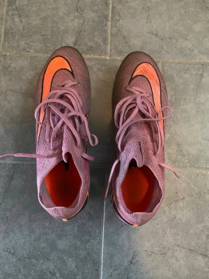 Lila-orange Nike fotbollsskor - Säljer ett par lila och orangea Nike elite modell fotbollsskor med högt skaft och snörning. Skorna har en tydlig Nike-logga på sidan och sulan är designad för bra grepp på planen. Materialet är syntetiskt och skon har en modern, strömlinjeformad look.