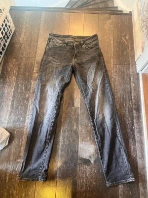 Svarta tapered jeans från Jack & Jones - Säljer ett par svarta tapered jeans från Jack & Jones med snygg tvättad look och klassiska fem fickor. Jeansen har gul söm på insidan av linningen och är tillverkade i ett mjukt denimtyg. Perfekta för dig som gillar en avslappnad men ändå stilren vibe. Skriv om ni har frågor!