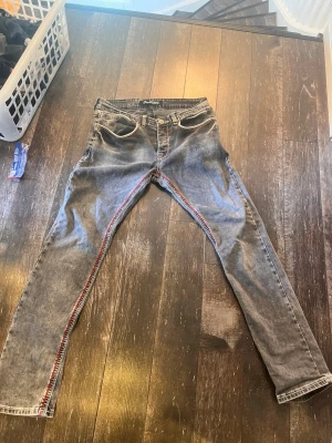 Grå jeans med röda sömmar OneRedox - Säljer ett par grå jeans från OneRedox med unika röda kontrastsömmar längs fickor och ben. Jeansen har fem fickor, bälteshällor och vit detaljsöm vid fickorna. Snygg tvättad look och raka ben. Perfekta för dig som vill sticka ut med detaljer! Skriv om ni har några frågor!