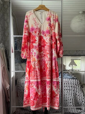 Lång blommig klänning med volang - Superfin långklänning i vitt med rosa och röda blommor, v-ringad och med långa ärmar. Klänningen har volangdetaljer och ett löst, flowy fall. Knytband i midjan. Aldrig använd, nypris 999 kr. 