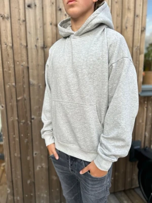 Ljusgrå hoodie med huva - En stilren ljusgrå hoodie med klassisk huva och stor magficka. Hoodien har ribbade muddar vid ärmslut och nederkant samt en avslappnad passform. Perfekt för en chill och enkel look. Materialet ser ut att vara mjuk bomullsblandning. Modellen är 179