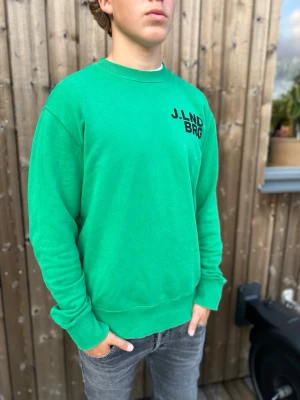 Grön sweatshirt J. Lindberg - Snygg grön sweatshirt med trycket J.LND BRG på bröstet. Tröjan är från J. Lindberg och har rund halsringning, ribbade muddar vid ärmslut och nederkant samt en avslappnad passform. Perfekt för en casual look och enkel att matcha med jeans. Modellen är 179