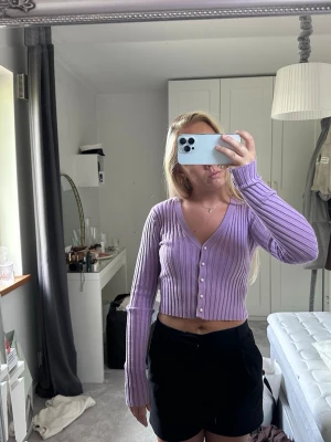 Ljuslila ribbad kofta med pärlknappar - Säljer en croppad, ljuslila ribbad kofta med långa ärmar och djup v-ringning. Koftan har snygga pärlknappar framtill och är figurnära i modellen. Perfekt för dig som gillar trendiga och feminina plagg. Från kollektionen AFJ X NA-KD.