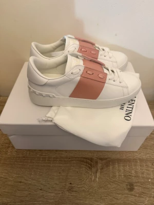Vita sneakers med rosa detalj - Snygga vita sneakers från Valentino med bred rosa rem över snörningen och coola nitar på hälen. Skorna har en chunky sula och är gjorda i slätt läder. Perfekta för dig som vill ha en clean men ändå unik look.