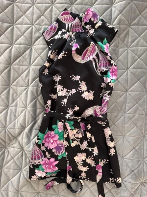 Blommig topp med blommor - Snygg svart topp med asiatiskt inspirerat blommönster i rosa, lila och grönt samt fågelmotiv. Klänningen är ärmlös, har hög krage och knytband i midjan. Perfekt för dig som vill sticka ut med en unik och färgglad look.