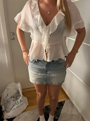 Vit prickig blus med volang - Säljer en söt, vit blus i transparent material med små prickar och volangdetaljer längs urringningen. Blusen har knyt framtill och korta ärmar som ger en luftig känsla. Perfekt för dig som gillar romantisk och trendig stil.