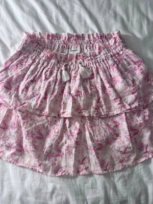 Rosa blommig volangkjol Kappahl - Söt kort kjol från Kappahl i ljusrosa med blommigt mönster i rosa toner. Kjolen har volangdetaljer och resår i midjan med dekorativa tofsar framtill. 
