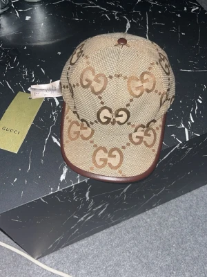 Beige keps med GG-mönster från Gucci - Snygg beige keps från Gucci med det klassiska GG-monogrammet i brunt över hela kepsen. Kepsen har en böjd skärm med mörkbrunt kantband och är tillverkad i ett slitstarkt tygmaterial. Perfekt för dig som vill ha en exklusiv och trendig accessoar.