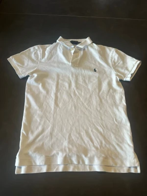 Vit pikétröja från Polo Ralph Lauren - Klassisk vit pikétröja från Polo Ralph Lauren i slim fit-modell. Tröjan har korta ärmar, krage och knappar framtill samt den ikoniska broderade loggan på bröstet. Tillverkad i mjuk bomull som känns skön mot huden.