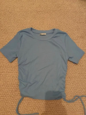 Ljusblå ribbad t-shirt med snörning - Säljer en ljusblå ribbad t-shirt från Zara med korta ärmar och rund hals. T-shirten har snörningsdetaljer i sidorna som ger en snygg twist. Perfekt för dig som gillar en enkel men trendig look. Man kan både använda den som en vanligt T-shirt eller dra till snörena vilket omvandlar tröjan till en cool topp.
