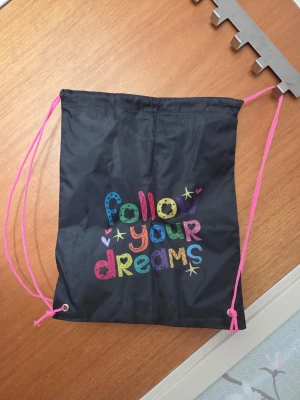 Svart gympapåse med färgglad text - Säljer en svart gympapåse med rosa snören och färgglad text på framsidan där det står 'follow your dreams' i olika mönster och färger. Perfekt för träning eller skola, lätt att bära på ryggen och enkel att stänga med dragsko.