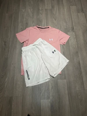 Under Armour träningsset rosa/vit - Säljer ett sportigt träningsset från Under Armour med en ljusrosa t-shirt och vita shorts. T-shirten har rund hals och korta ärmar, medan shortsen har resår i midjan och logga på benet. Perfekt för gymmet eller löpning.