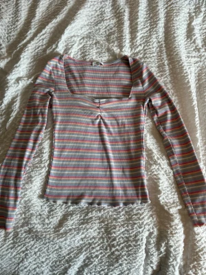 Randig långärmad topp med rynk - Snygg långärmad topp med smala ränder i rosa, blått, gult och vitt. Toppen har fyrkantig urringning och en rynkad detalj framtill. Materialet är ribbat och stretchigt för en bekväm passform.