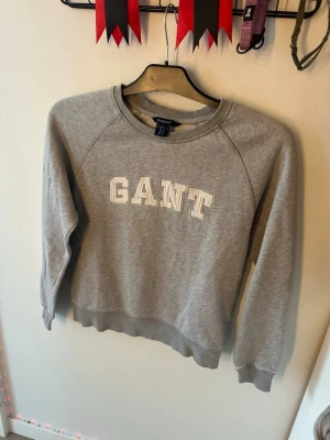 Grå sweatshirt från GANT - Klassisk grå sweatshirt från GANT med broderad logga i vitt på bröstet. Tröjan har rund halsringning, ribbade muddar vid ärmslut och nederkant samt en avslappnad passform. Perfekt för en casual och sportig stil.