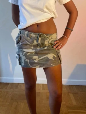 Camouflage kortkjol med fickor - Säljer en snygg camouflage kortkjol med stora fickor och dubbla knappar i midjan. Kjolen har en cool militärstil och är perfekt för dig som vill sticka ut. Tillverkad i bomull för en bekväm känsla och passar till många olika looks. Den står storlek 38, men sitter som 36! Första bilden är lånad!