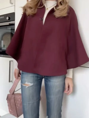 Vinröd cape-kappa med knapp - Stilren vinröd cape-kappa med vid passform och hög krage. Kappan har en knapp framtill och två fickor. Perfekt för lager-på-lager och ger en trendig siluett. Materialet ser ut att vara ullblandning för extra värme.