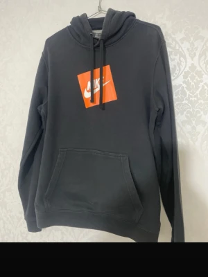 Svart Nike hoodie med orange logga - Säljer en svart hoodie från Nike med stor orange fyrkantig logga på bröstet och vit Nike-text. Hoodien har huva med dragsko och en stor magficka. Perfekt för dig som gillar streetwear och vill ha en enkel men snygg look.