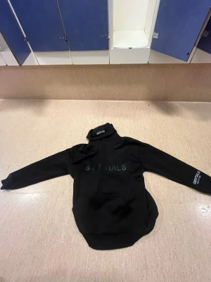 Svart Essentials hoodie med tryck - Säljer en svart hoodie från Essentials med stor logga på bröstet och tryck på ärmen. Hoodien har en klassisk huva och magficka. Perfekt för dig som gillar streetwear och vill ha en stilren look.
