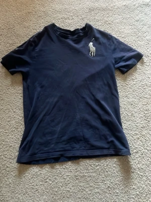 Mörkblå t-shirt med stor logga - Snygg mörkblå t-shirt med en stor, vit broderad logga på bröstet. Klassisk rund hals och korta ärmar. Perfekt till jeans eller shorts för en clean och stilren look.