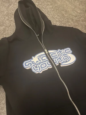 Galactic Svart Hoodie Med Full zip-up - Säljer en svart hoodie med dragkedjan som dras upp i luvan också, med en blå riktigt fet figur i baksidan.