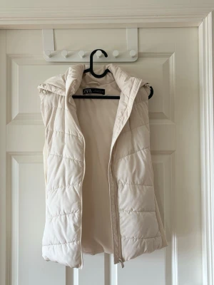 Beige dunväst med huva från ZARA - Stilren beige dunväst från ZARA med quiltad design och mysig huva. Västen har dragkedja framtill och är lätt vadderad för extra värme. Perfekt att slänga över en hoodie eller tröja för en trendig lager-på-lager-look.