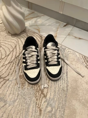 Svarta och vita sneakers med ränder - Snygga sneakers i svart och vitt med klassiska ränder på sidorna och perforerad tå. Skorna har vita snören och svart sula. Perfekt för dig som gillar en sportig och stilren look. Materialet är en mix av textil och syntet.