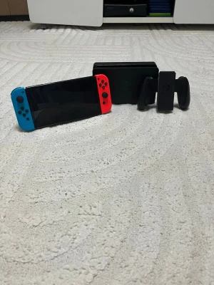 Nintendo Switch med neonröda och blå Joy-Con - Nintendo Switch spelkonsol med neonröda och blå Joy-Con-kontroller. Konsolen är svart och har en rektangulär form. Dockningsstation och Joy-Con grip ingår. Perfekt för gaming både hemma och på språng. Tyvär så är en av kablarna är borta så de måste ni köpa själva 