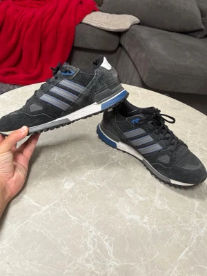 Adidas ZX 750 skor - Snygga Adidas ZX 750 sneakers i svart mocka och mesh med blå och grå detaljer. Skorna har de klassiska tre ränderna på sidan, vit och blå sula samt ZX 750-märke på hälen. Sjukt trendiga just nu och passar perfekt som vardagliga skor men även vid finare tillställningar. 