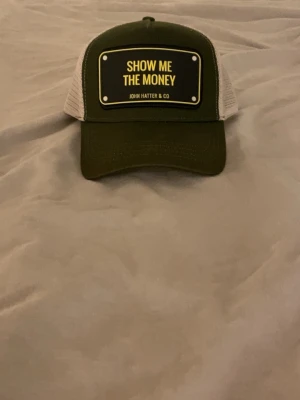 Grön och vit truckerkeps John Hatter & Co - Snygg truckerkeps i grön med vit mesh och justerbar snapback. Framtill finns en gul textplatta med texten 'SHOW ME THE MONEY'. Perfekt för dig som vill sticka ut med en cool accessoar.
