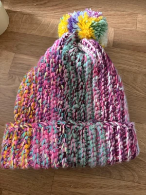Flerfärgad stickad mössa med boll - Säljer en riktigt färgglad och chunky stickad mössa med uppvikt kant och stor garnboll på toppen. Mössan har inslag av rosa, lila, gult, blått och vitt i garnet, vilket ger en unik och lekfull look. Perfekt för dig som vill sticka ut i vinter och hålla dig varm.