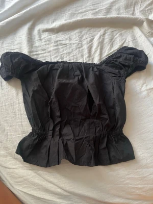 Svart offshoulder topp med puffärm - Trendig svart offshoulder topp med puffiga korta ärmar och smockad midja. Toppen har en lätt volang nedtill och är perfekt för dig som gillar en feminin och cool stil. Materialet känns som bomull och toppen är enkel att matcha med både jeans och kjol.