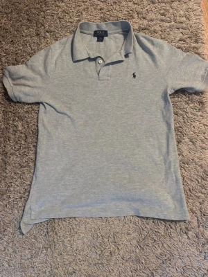 Piké  Polo Ralph Lauren - Klassisk ljusgrå pikétröja från Polo Ralph Lauren med korta ärmar och liten broderad logga på.Perfekt till en avslappnad men ändå stilren look. är helt ny och har aldrig andvänt den då den var för stor för mig 