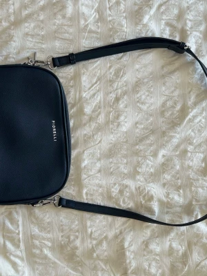 Mörkblå axelväska från Fiorelli - Snygg marinblå axelväska från Fiorelli i stilren design. Väskan har två stora fack med silverfärgade dragkedjor och justerbar axelrem. Tillverkad i slätt konstläder och har diskret logga på framsidan. Perfekt för dig som gillar minimalistisk stil.
