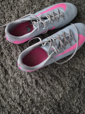 Ljusgrå fotbollsskor med rosa detaljer - Säljer ett par snygga fotbollsskor i ljusgrått med skarpa rosa detaljer och snörning. Skorna har en låg profil och är designade för bra grepp på planen. Insidan är fodrad i rosa och ovandelen är i ett syntetmaterial som ger bra bollkontroll.