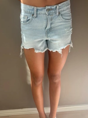 Ljusblå jeansshorts med fransig kant - Snygga ljusblå jeansshorts med fransig och rå kant nertill. Klassisk femficksmodell med knapp och dragkedja framtill. Shortsen har slitna detaljer och är perfekta för sommaren. Passformen är normal och midjan är midwaist.