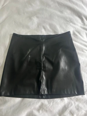 Svart kort kjol i skinnimitation - Säljer en svart kort kjol från H&M Divided i skinnimitation. Kjolen har en clean och stilren look med rak passform och är perfekt för att skapa en edgy outfit. Materialet är mjukt och följsamt syntetmaterial.