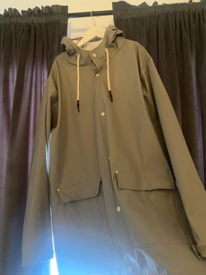 Grå regnjacka med huva - Snygg beige regnjacka från lager 157 med huva och vita dragsnören. Jackan har tryckknappar framtill och två stora fickor med lock. Lång modell som skyddar bra mot regn och vind. Perfekt för blöta dagar och enkel att matcha med olika outfits. Jackan är i mycket bra skick! Priset är förhandlingsbart!
