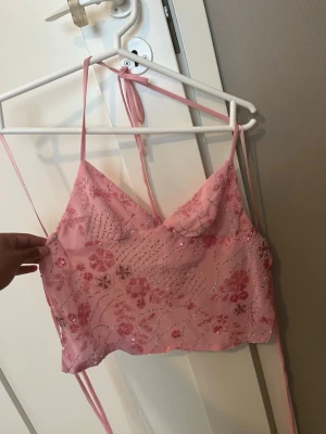 Rosa blommig sequin topp - Söt rosa croppad linnetopp med tunna axelband och blommigt mönster i paljetter. Toppen har v-ringning och knytband i ryggen för justerbar passform. Perfekt för sommarens varma dagar och ger en romantisk vibe. Den är XXS men passar för mig som är S! Jag har bara använt den en gång senn jag köpte den!