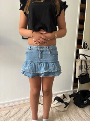 Blå volang jeanskjol - Säljer en ljusblå jeanskjol med volangkant och klassisk femficksdesign. Kjolen har en kort passform och är gjord i denim med bälteshällor och knappgylf. Perfekt för dig som vill ha en trendig och avslappnad look.