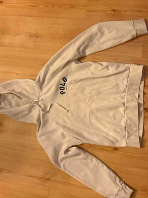 Vit hoodie från Polo Ralph Lauren - Säljer en stilren vit hoodie från Polo Ralph Lauren med svart logga på bröstet. Hoodien har huva med dragsko och en stor magficka. Perfekt för dig som gillar klassisk streetwear och vill ha något bekvämt och snyggt.