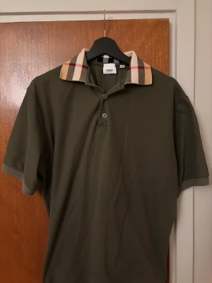 Burberry piké  - Snygg olivgrön burberry pikétröja med klassisk rutig krage i beige, svart och rött. Tröjan har korta ärmar, knäppning framtill. Perfekt för dig som gillar stilrena detaljer och vill sticka ut lite extra.