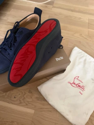 Mörkblå sneakers från Louboutin - Snygga mörkblå sneakers från Christian Louboutin med klassisk röd sula och diskret logga på insidan. Skorna har en låg profil, rund tå och är tillverkade i mocka med matchande mörkblå snören. Perfekta för dig som vill sticka ut med exklusiv stil.