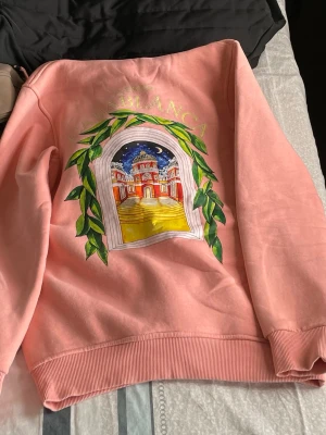 Rosa hoodie med tryck från Casablanca - Säljer en rosa hoodie från Casablanca med stor färgglad grafik på ryggen och mindre tryck framtill. Motivet visar en byggnad i solnedgång och gröna blad runt ett fönster. Hoodien har huva med dragsko och magficka. Mjuk bomullskvalitet och ribbade muddar.