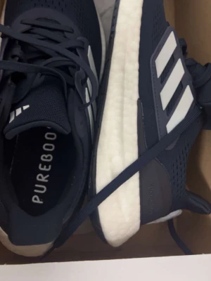 Adidas Pureboost svarta sneakers - Säljer ett par svarta Adidas Pureboost sneakers med vita detaljer och klassiska tre ränder på sidan. Skorna har en tjock, vit Boost-sula för extra komfort och ovandel i mesh för bra andningsförmåga. Perfekta för dig som gillar sportig stil.