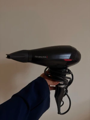 Svart hårfön från Remington - Säljer en svart hårfön från Remington med slimmad design och detaljer i rött. Hårfönen har ett smalt munstycke och lång sladd som är enkel att vira ihop. Perfekt för styling av håret hemma eller på resan.